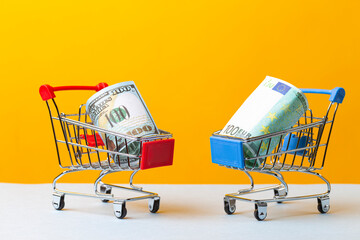 Savings Ideas. Two Shopping Trolleys Carts With US Dollars and Euro Currency Banknotes Over Trendy Yellow Background.