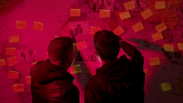 Two unrecognizable young men looking at wall map with strings, stickers and photos, investigating connections, talking and pointing. Back view. Low key. Investigation, project concept