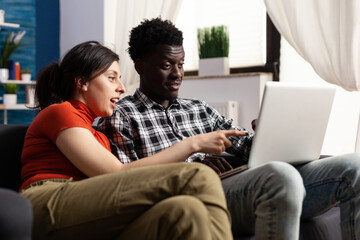 Joyful young multiethnic couple looking at computer screen while sitting on couch in the living room. People enjoying being together by casual websurfing and watch interesting videos on internet.