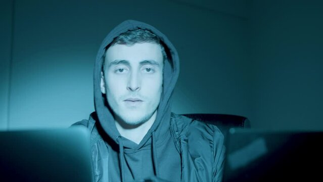 Young Caucasian man sitting at night in front of laptop screen and hacking. Man looking at camera, then intertwining fingers and lifting his eyebrows. Hacking, cyber crime concept