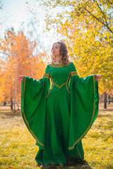 A beautiful young woman in a historical green dress stands in the autumn forest. Medieval queen among dry leaves wearing a crown.