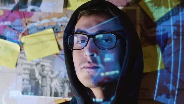 Portrait shot of Caucasian man in hood against investigation wall map. Young hacker turning head and looking at camera while projector video image reflecting on his face. Hacking, youth concept