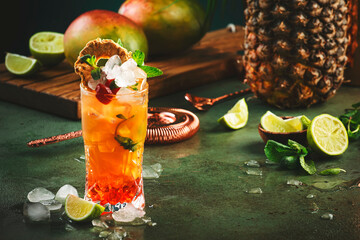 Alcoholic cocktail with vodka, pineapple juice, mango, red syrup and ice. Long drink or summer cold mocktail. Tropical dark background with palm leaves and exotic fruits
