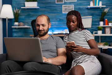 Happy caucasian man websurfing on laptop while african american woman holding smartphone and looking at computer screen. Couple sitting on sofa in living room checking bills and financial issues on