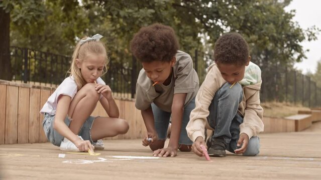 Slowmo of pretty 10 year old Caucasian girl and two African American boys eating lollipops and drawing with chalk outdoors in summer