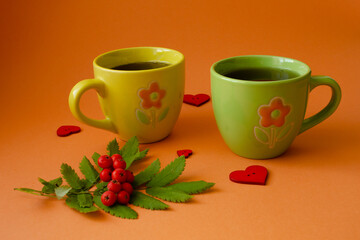 Yellow and green mugs with ripe rowan berries and red hearts on orange background