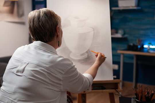 Over shoulder view of older woman drawing orginal masterpiece using sharp pencil on paper sketching vase model in home art studio. Portrait of elderly artist sketching still life in workshop.