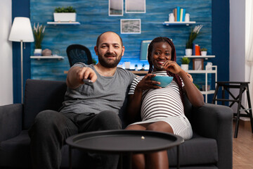 Happy interracial couple watching TV show while sharing a bowl of popcorn sitting on sofa. Smiling future parents expecting baby resting at home while having a leisure time together.