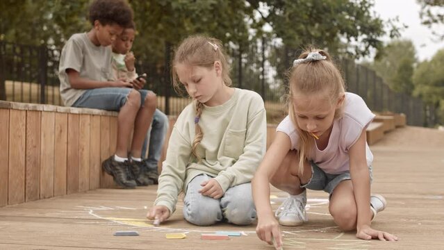 Slowmo of two pretty 10 year old Caucasian girls drawing with chalk on wooden planks while two African American boys playing games on smartphone in background, spending summertime outdoors in park