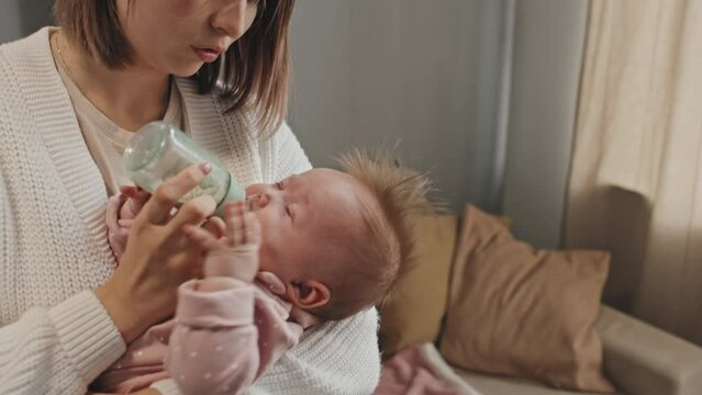 Tilting up of young Biracial parent standing in living room, holding crying blue-eyed daughter, giving her water in feeding bottle, rocking infant at daytime