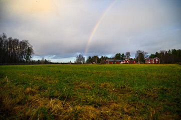 A cloudy landscape view of open field filled with grass with cottages and rainbow in the sky in a rainy day during a outdoor visit