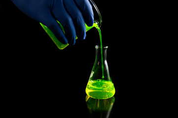 A research scientist experimenting with a green fluorescent droplets in a glass conical flask in dark biomedical laboratory for health care medicine development.