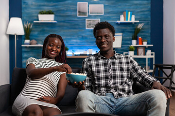 Smiling young african american couple sharing a bowl of popcorn while watching a television comedy program. Happy pregnant woman enjoying eating snacks sitting on couch while watching a movie in