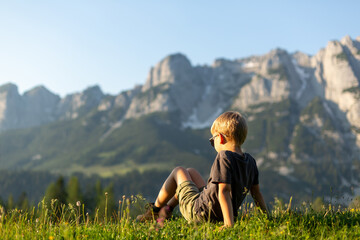 Naklejka na ściany i meble A boy in hiking boots sitting on the grass in mountains. Outdoor activities concept. Austrian Alps
