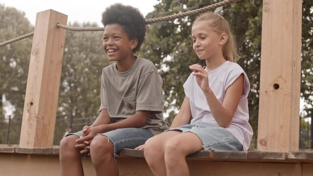 Slowmo of African American boy and Caucasian girl sitting outdoors on wooden planks with hanging feet, talking and holding hands