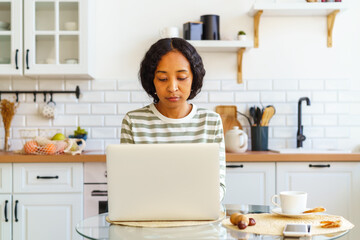 Focused African-American female working from kitchen on laptop. Concept of home remote office