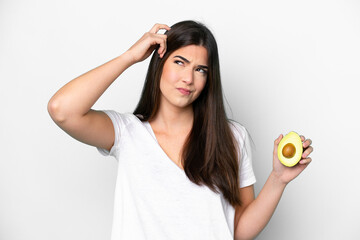 Young Brazilian woman holding an avocado isolated on white background having doubts and with...