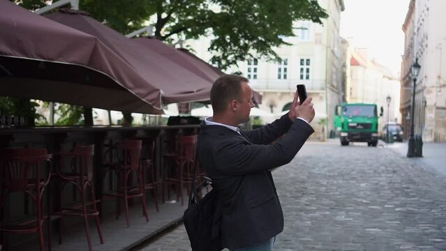 Side view of man walking on ancient street with beautiful medieval facade, male tourist taking pictures and recording video on mobile phone.