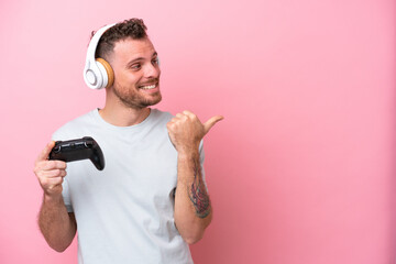 Young Brazilian man playing with video game controller isolated on pink background pointing to the side to present a product