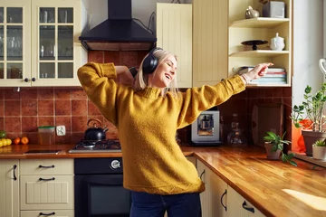 Gordijnen Joyful mid adult woman dancing in kitchen listening music on wireless headphones © baranq