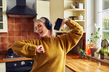 Joyful mature woman dancing in kitchen while listening music on headphones