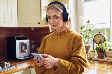 Beautiful mature woman using mobile phone listening music on wireless headphones in kitchen