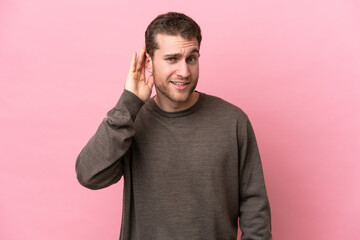 Young caucasian man isolated on pink background listening to something by putting hand on the ear