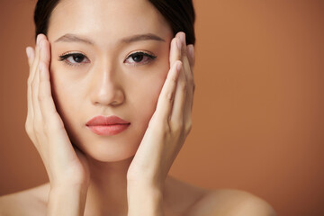 Face of serious young Vietnamese woman touching her flawless soft skin and looking at camera