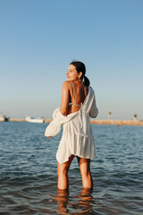 A girl with a gorgeous figure at sea at sunset. Sexy happy smiling girl in a white dress on a wild beach. Girl in the water, catches the rays of the sun