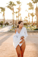 Fototapeta premium Portrait of a beautiful young lady in a white bikini. A model poses among exotic palm trees against a boho backdrop. Stylish photo shoot on vacation.
