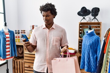 African man with curly hair holding shopping bags using smartphone in shock face, looking skeptical...