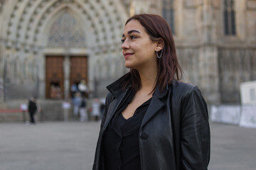 headshot portrait with copy space of a likable latin woman dressed with black clothes who is standing on a street of Barcelona where there is a church that is behind her