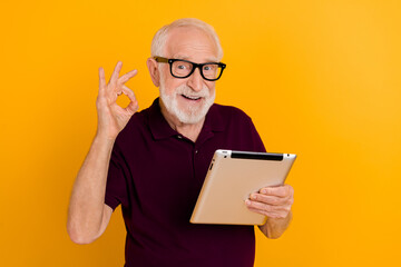 Photo of cheerful mature man show fingers okey symbol approve good tablet app isolated over yellow...