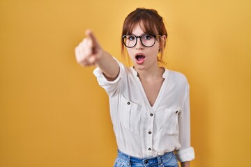 Young beautiful woman wearing casual shirt over yellow background pointing with finger surprised ahead, open mouth amazed expression, something on the front