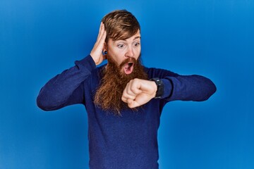Redhead man with long beard wearing casual blue sweater over blue background looking at the watch...