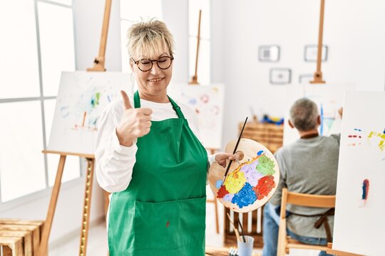 Middle age caucasian woman drawing canvas at art studio smiling happy and positive, thumb up doing excellent and approval sign
