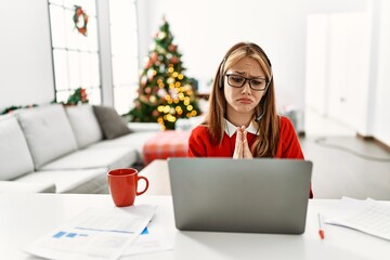 Young caucasian girl sitting on the table working using laptop by christmas tree begging and...