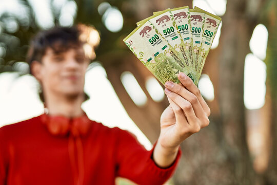 Young hispanic man using headphones holding argentinian pesos at the city