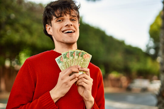 Young hispanic man smiling happy holding argentinian pesos banknotes at the city
