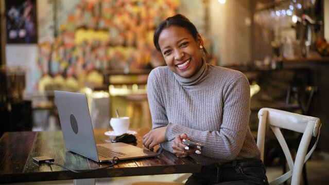African American Business woman looking at camera while working with laptop at cafe bar restaurant. Female remote worker using computer sitting in a table and drinking coffee indoors 
