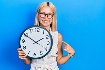 Beautiful blonde woman holding big clock smiling happy pointing with hand and finger
