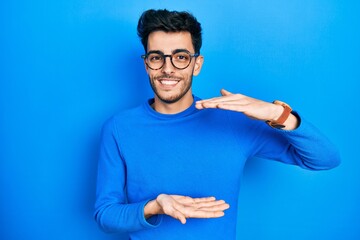 Young hispanic man wearing casual clothes and glasses gesturing with hands showing big and large size sign, measure symbol. smiling looking at the camera. measuring concept.