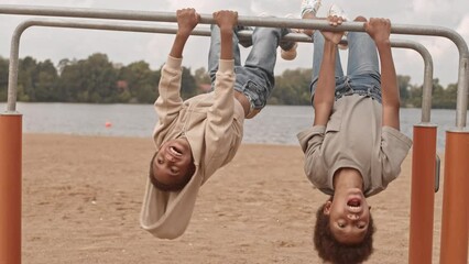 Slowmo of two cute African American sibling boys laughing while hanging upside down in gymnastic bar at playground on sandy beach near lake - Powered by Adobe