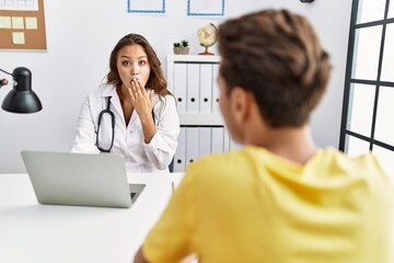 Young doctor woman with patient at the clinic covering mouth with hand, shocked and afraid for...
