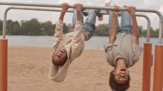 Slowmo of two cute African American sibling boys laughing while hanging upside down in gymnastic bar at playground on sandy beach near lake