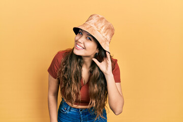 Young hispanic girl wearing casual clothes and hat smiling with hand over ear listening an hearing to rumor or gossip. deafness concept.