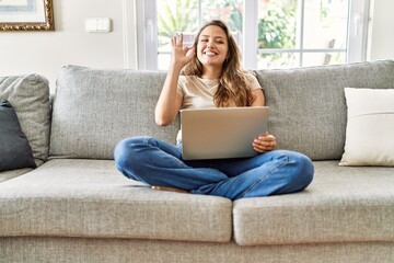 Beautiful young brunette woman sitting on the sofa using computer laptop at home smiling positive...