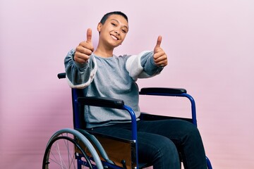 Beautiful hispanic woman with short hair sitting on wheelchair approving doing positive gesture with hand, thumbs up smiling and happy for success. winner gesture.