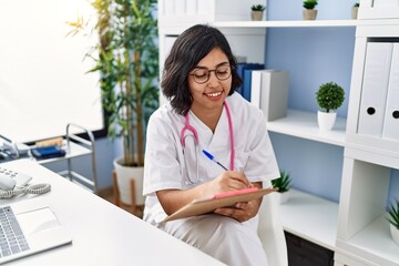 Young latin woman wearing doctor uniform writing on clipboard at clinic