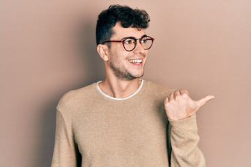 Young hispanic man wearing casual clothes and glasses smiling with happy face looking and pointing to the side with thumb up.